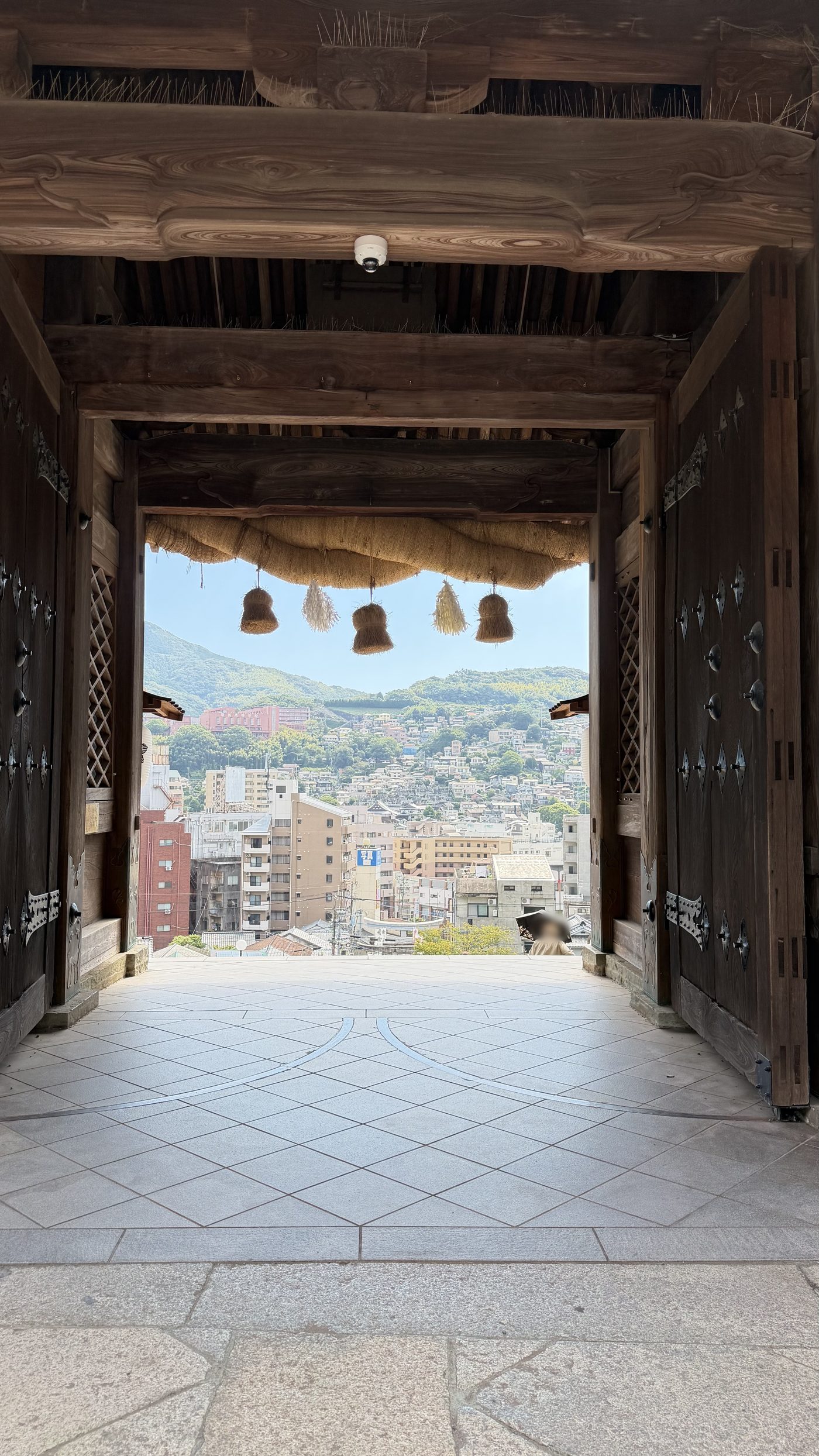 木造の門の向こうに、長崎の街並みと山並みが広がる風景